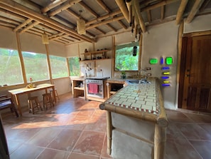 A rustic kitchen with a wooden ceiling, a stove, a sink, and a window overlooking greenery.