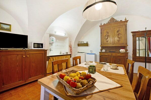 A dining area with a wooden table, chairs, a fruit basket, a television, and a kitchen in the background.