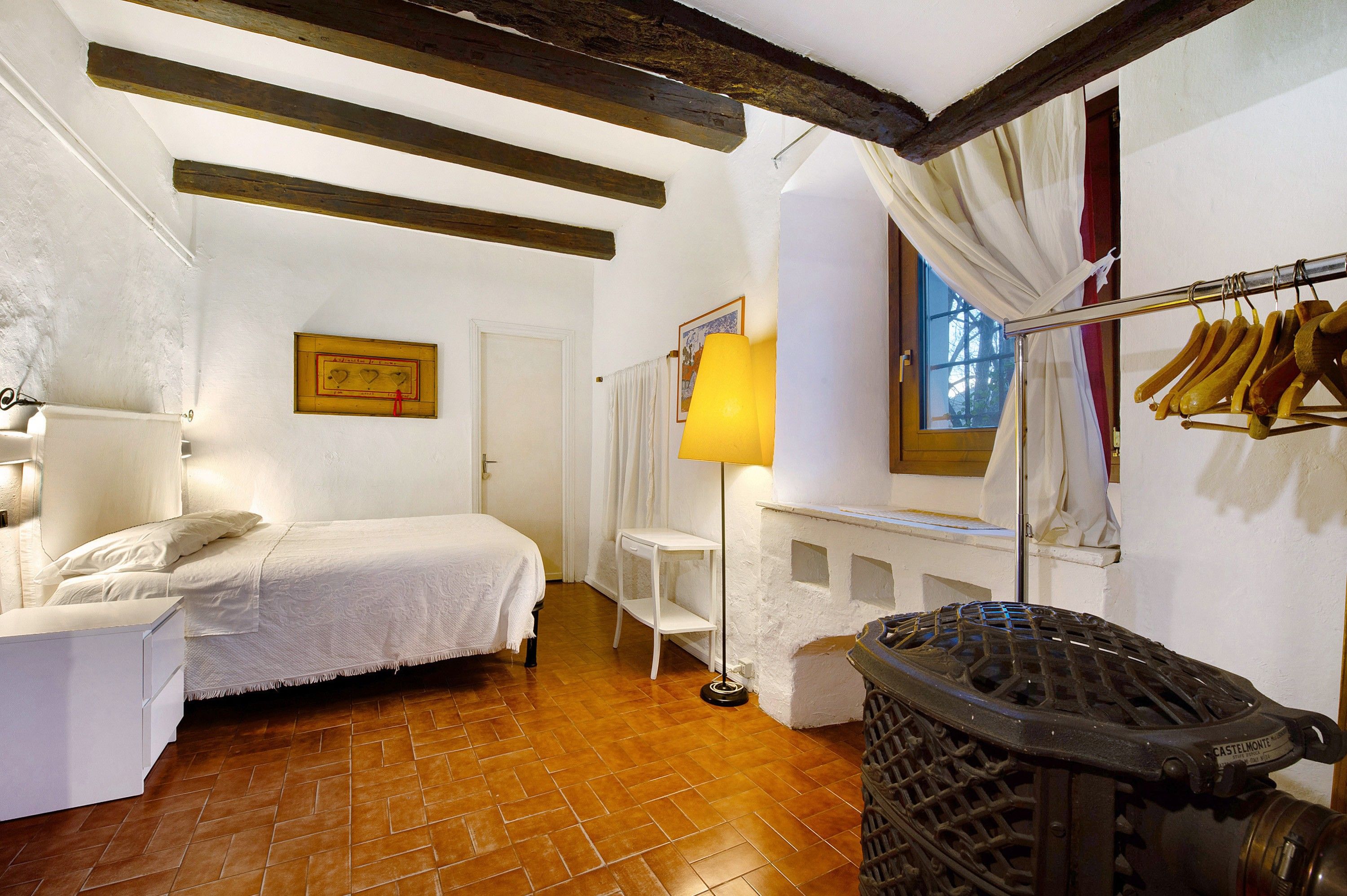 Une chambre à coucher comprenant un lit, une table de chevet, une lampe, un radiateur, une fenêtre avec des rideaux et un porte-manteau.