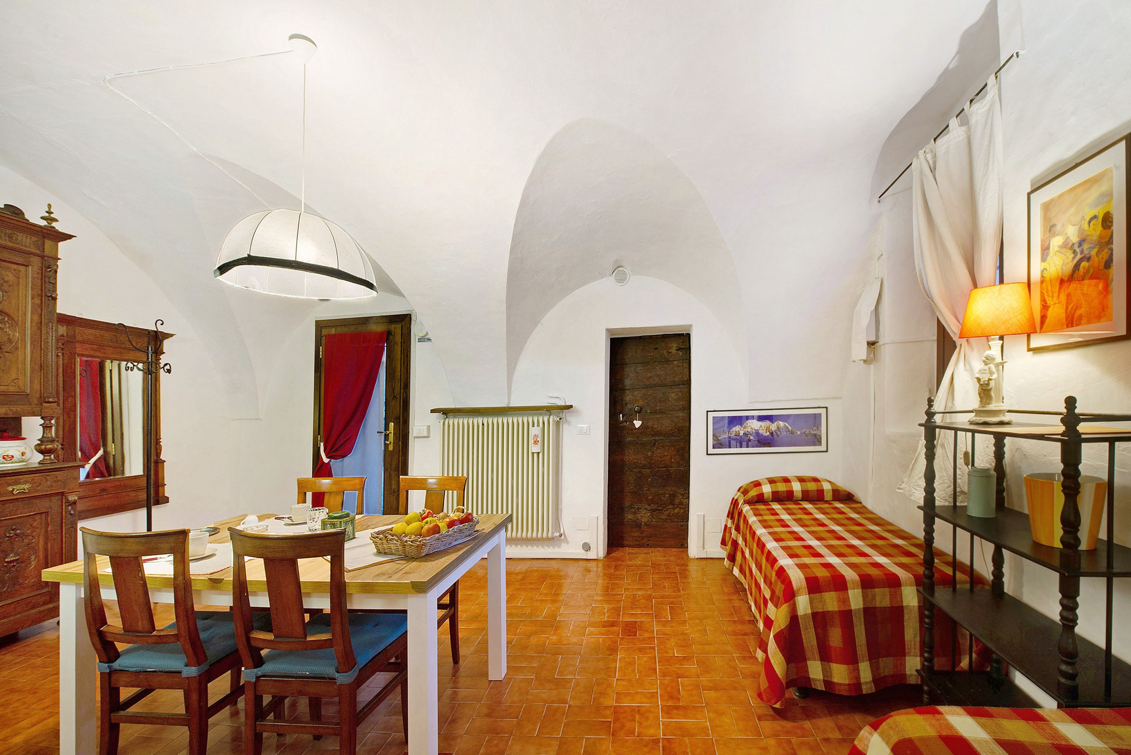Una habitación con una mesa de comedor, sillas, una cama con colcha a cuadros, un radiador, una lámpara de pared y un cuadro enmarcado.