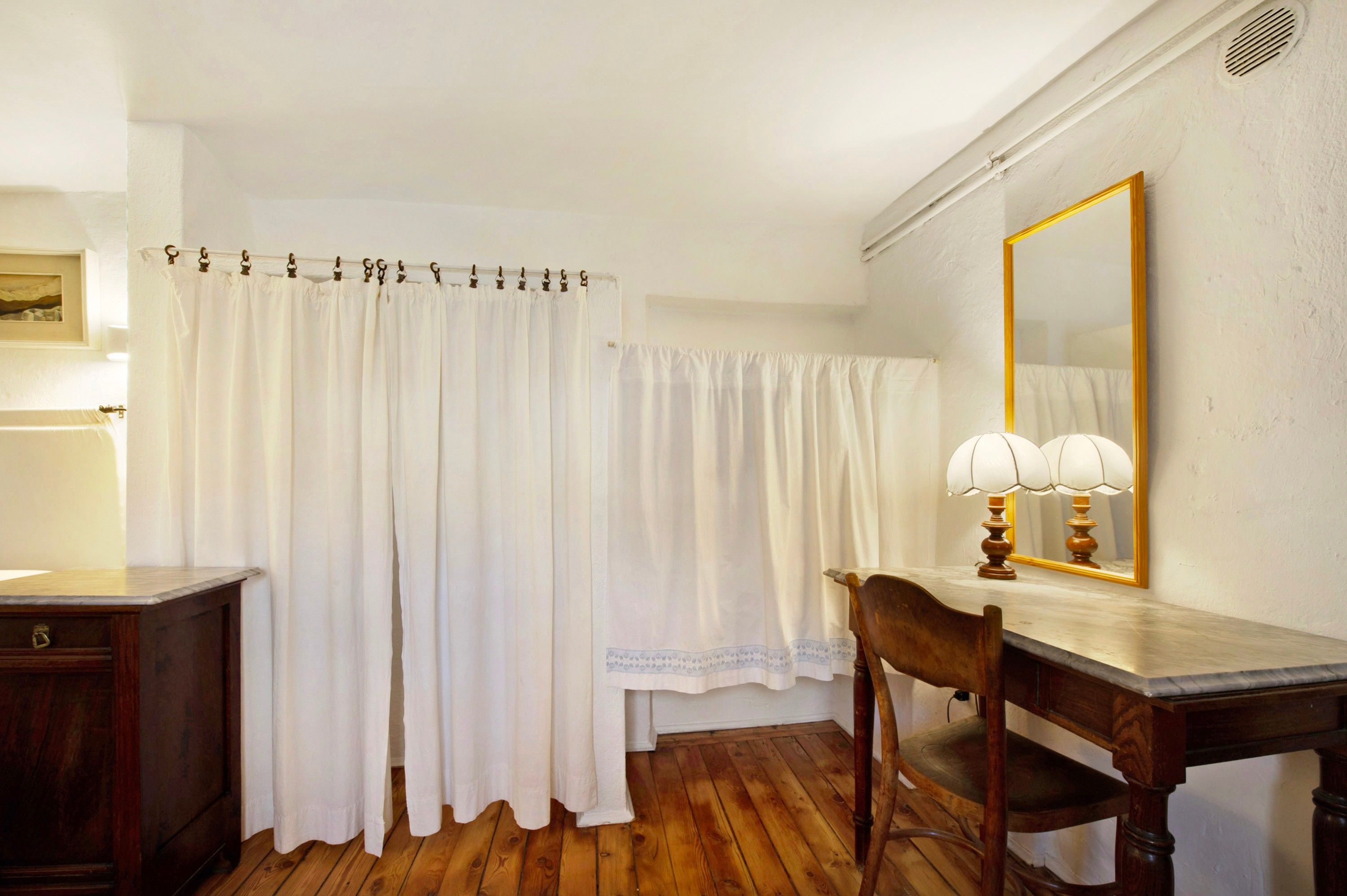 Une pièce avec un plancher en bois, un bureau en bois avec une lampe, une chaise, un rideau blanc et un miroir au mur.