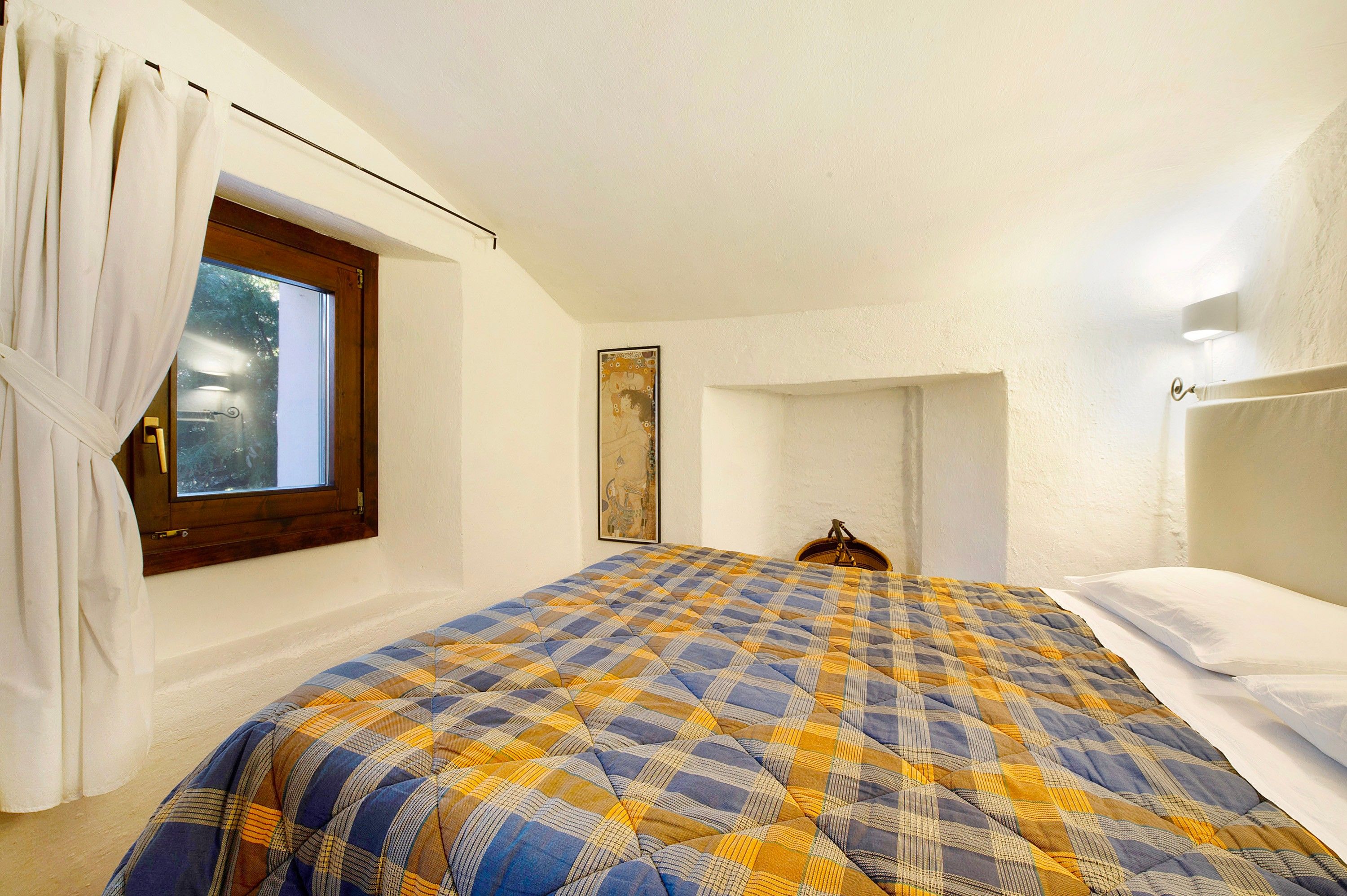 Une chambre avec une couvre-lit à carreaux, une fenêtre avec des rideaux, une tête de lit en bois et un tableau accroché au mur.