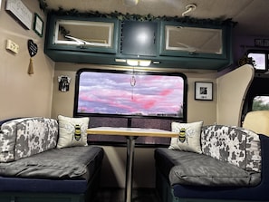 A cozy interior with a bed, a table, and a large window with a pink sky view.