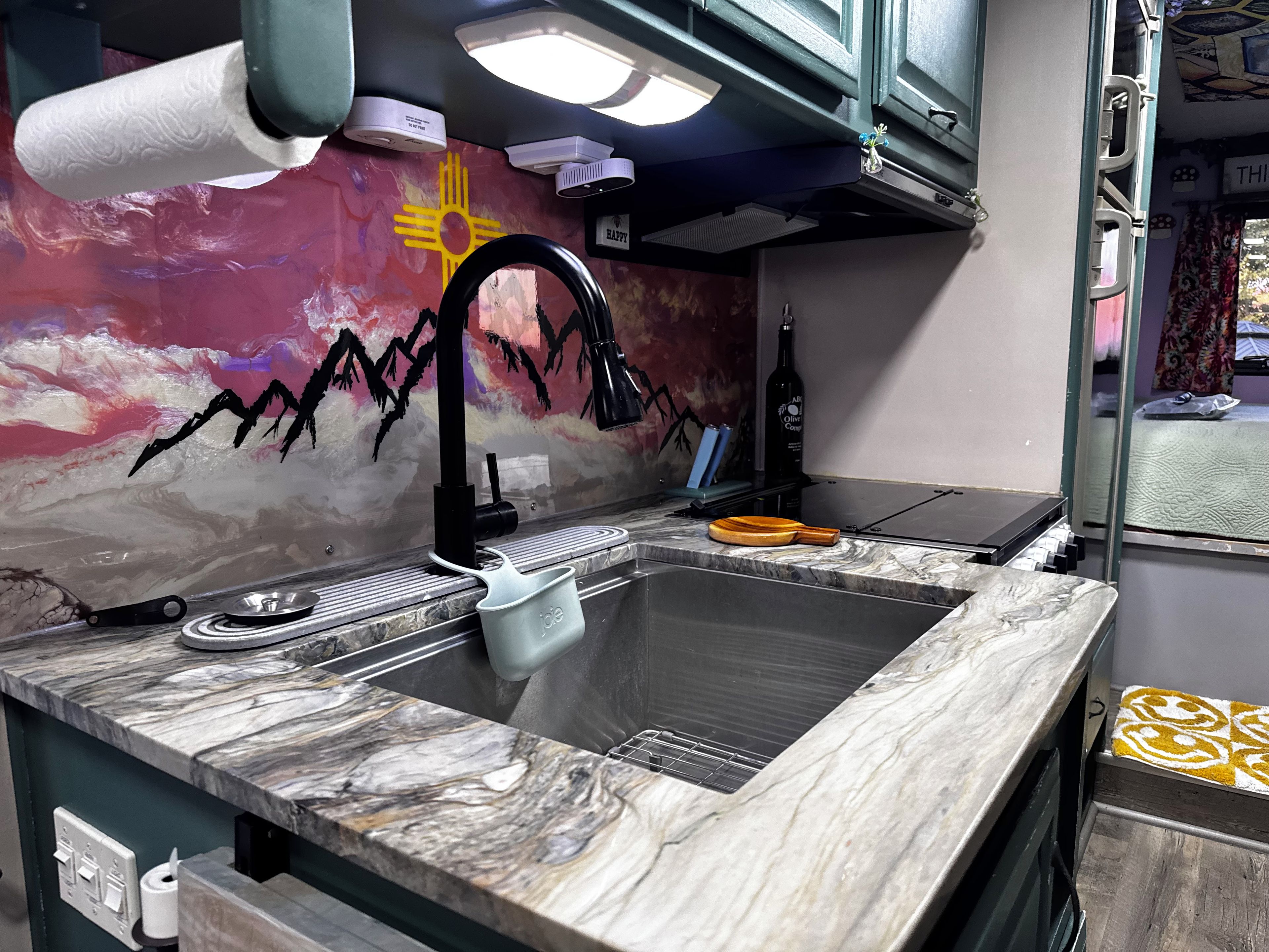 A kitchen with a marble countertop, a sink, a faucet, a bottle of olive oil, and a wall with a mural.