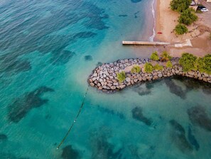An aerial view of a coastal area with a rocky breakwater, clear turquoise water, and a sandy beach with sparse vegetation. - Dorado Cove | Modern Pool & Beach Escape (Dorado)