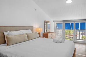 A hotel room with a large bed, a nightstand with a lamp, a wooden dresser, and a balcony with blue shutters. - Dorado Cove | Modern Pool & Beach Escape (Dorado)