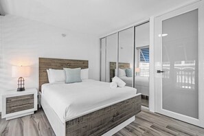 A modern bedroom with a large bed, wooden headboard, white bedding, a nightstand with a lamp, and a sliding glass door leading to a balcony. - Dorado Cove | Modern Pool & Beach Escape (Dorado)
