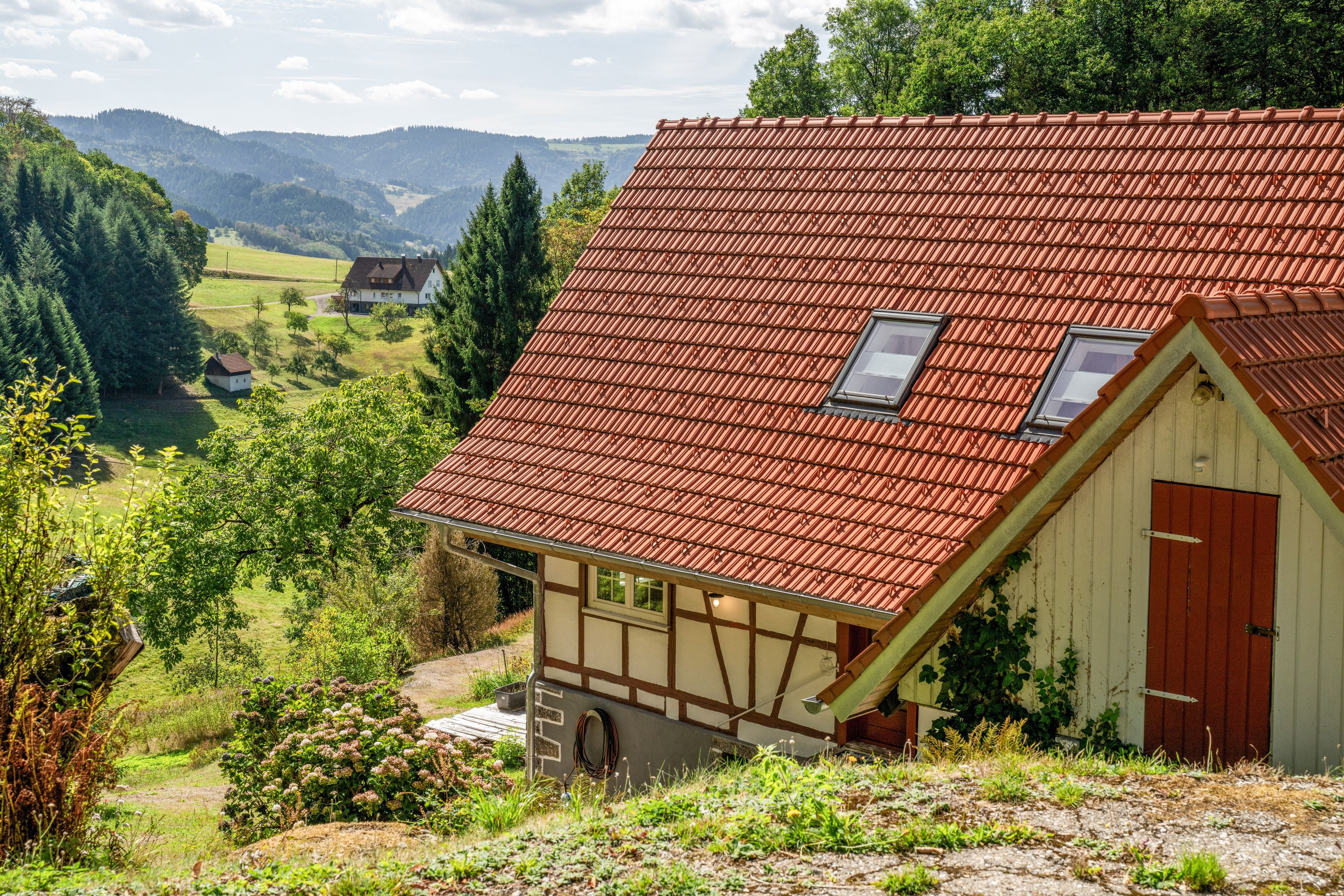 A traditional chalet with a sloped red roof, a wooden door, and a view of a valley.