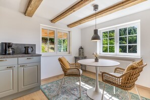 Eine moderne Küche mit Essbereich, inklusive eines runden Tisches, Korbstühle und Blick ins Grüne durch große Fenster.