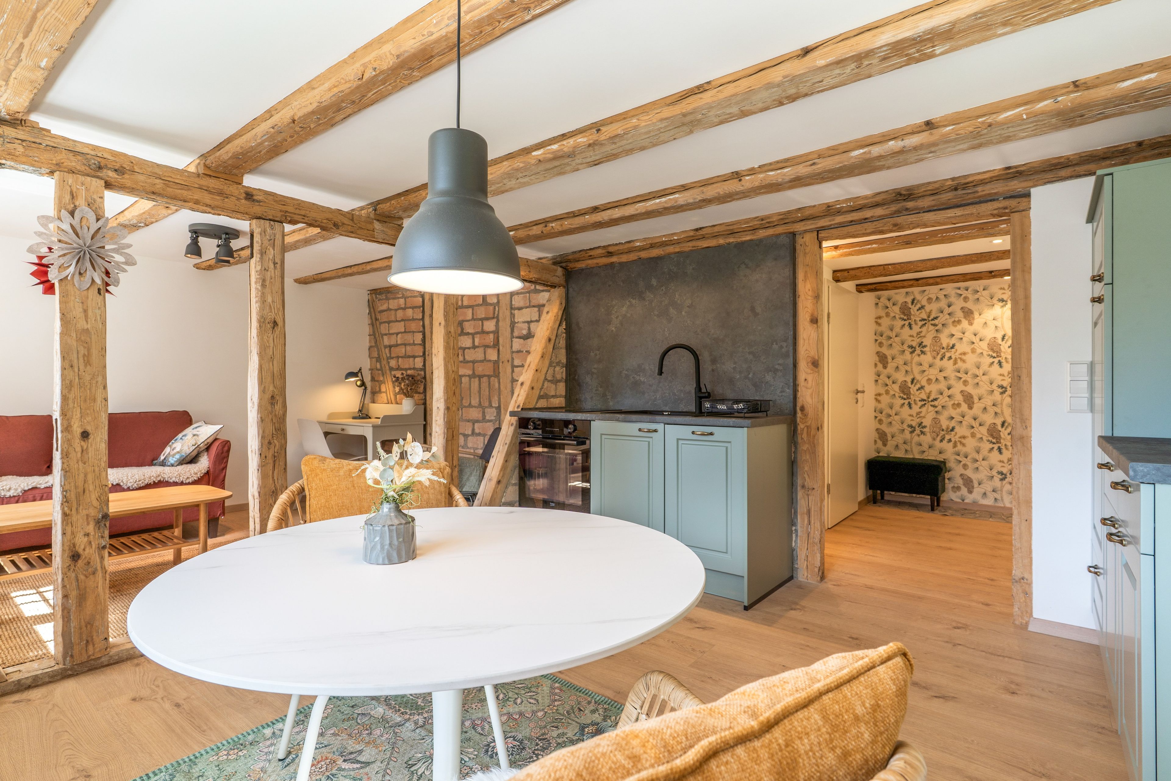 Un intérieur chaleureux avec une table de salle à manger ronde, des poutres en bois, une cuisine aux armoires bleues et un papier peint à motifs.