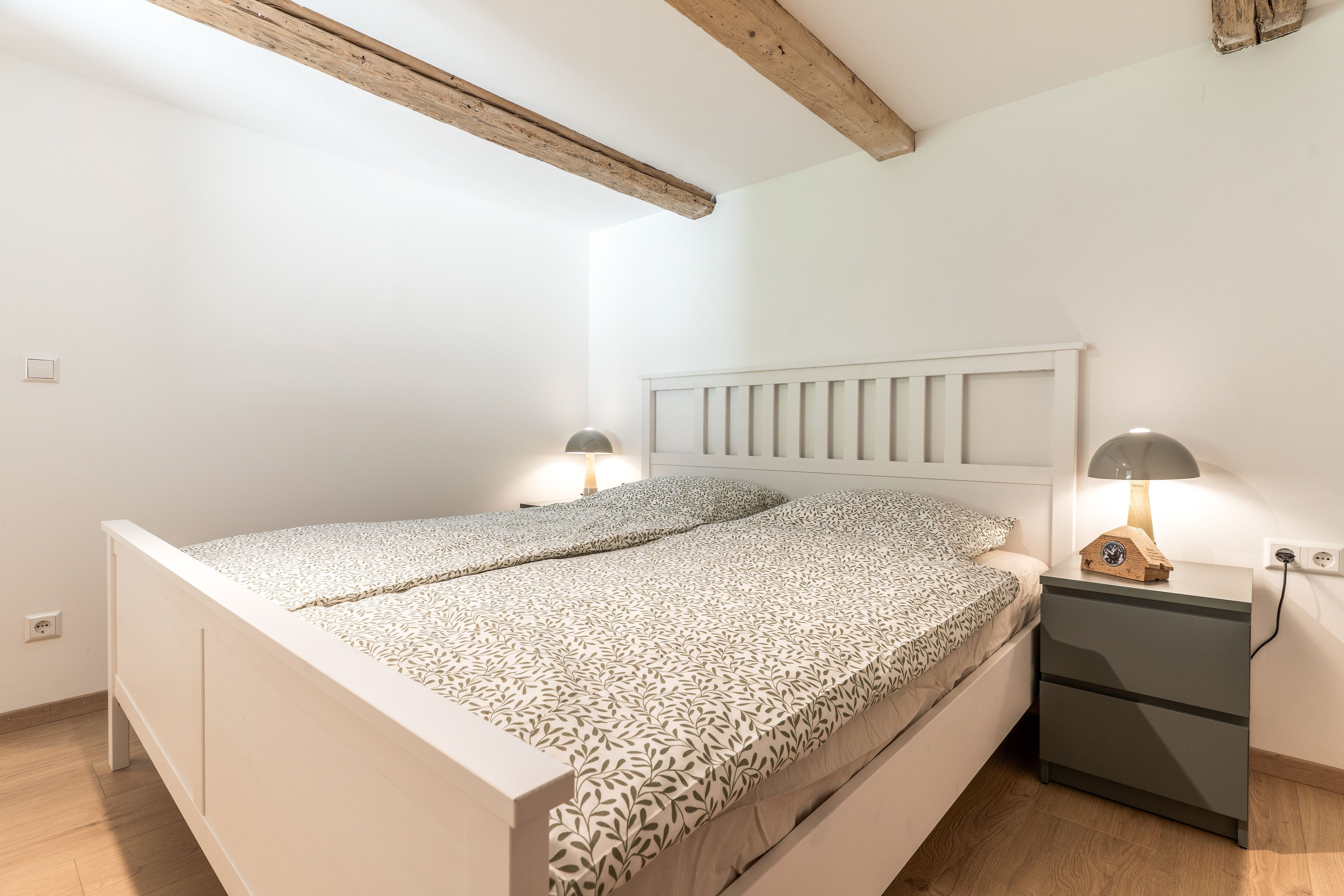 Ein Schlafzimmer mit einem weißen Bett, zwei Nachttischen mit Lampen, einem Holzbalken an der Decke und einer weißen Wand.