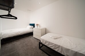 A bedroom with two single beds, a bedside table with a lamp, and a hanging coat rack. - Finest Retreats | Westwell Hall (Ilfracombe)