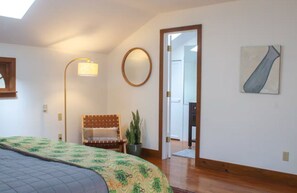 A bedroom with a bed, a bedside lamp, a mirror, a plant, and a door leading to another room. - LIBRARY HOUSE IN THE WOODS NEAR CHATHAM (Chatham)