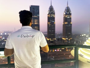 Een persoon in een wit shirt met een hashtag en een logo, kijkt uit over een stadsgezicht met twee opvallende wolkenkrabbers.