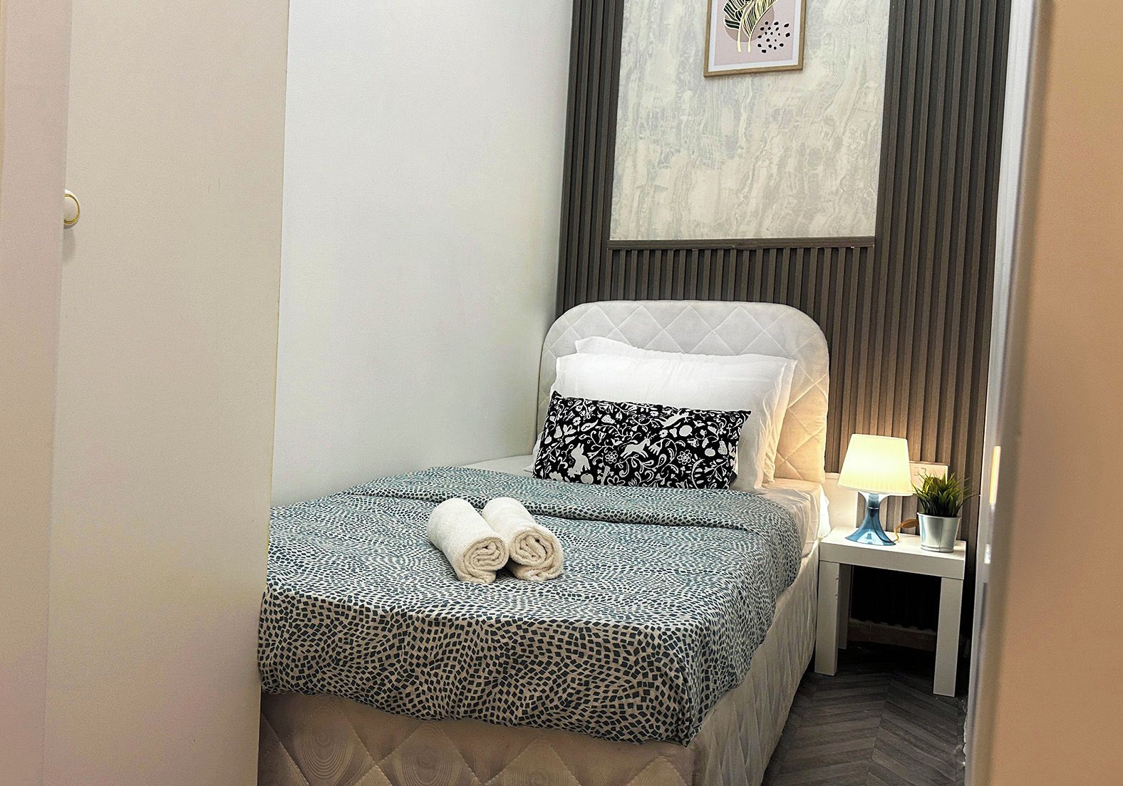 Een net opgemaakt bed met behangpatroon, een bedlampje en een kleine potplant op een nachtkastje.