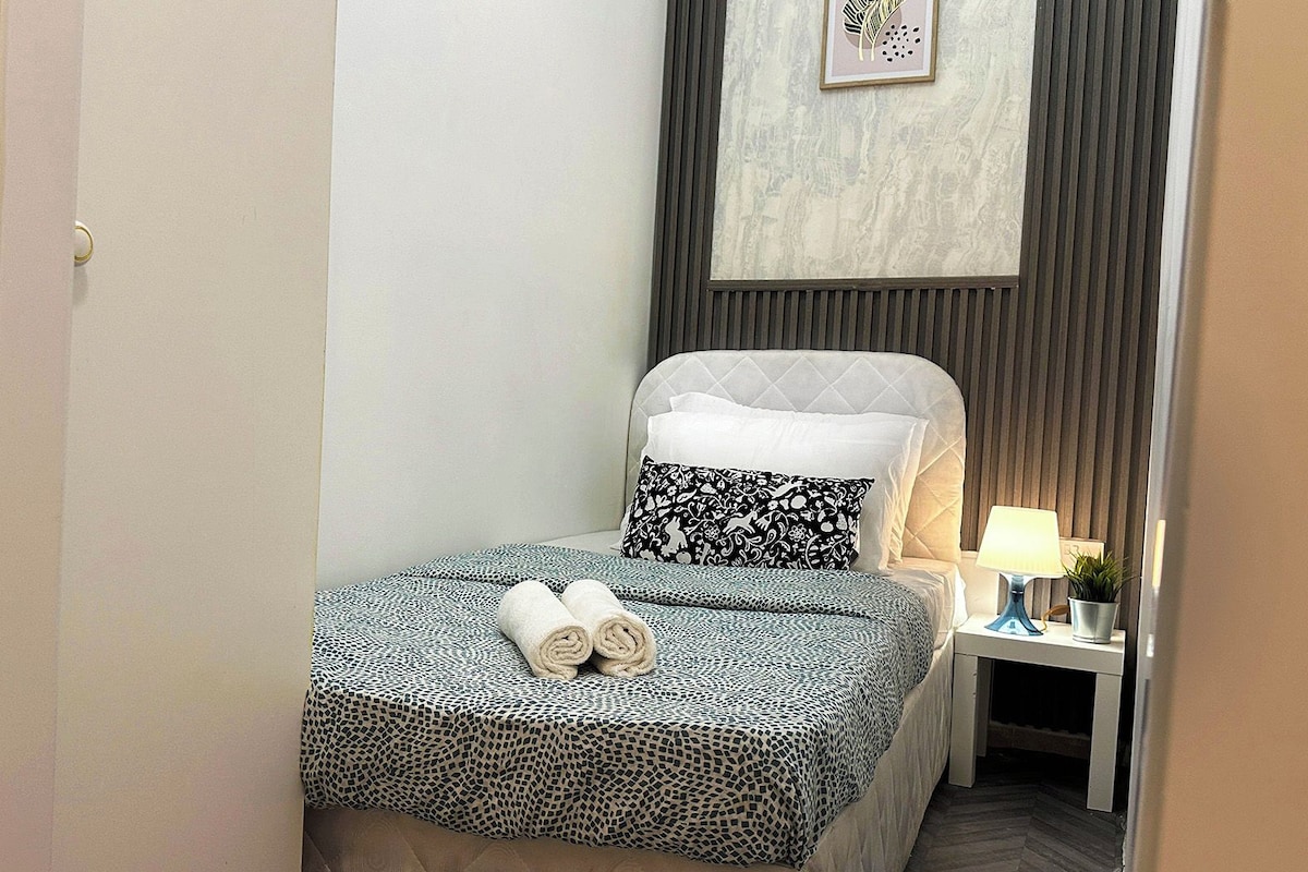 Een net opgemaakt bed met behangpatroon, een bedlampje en een kleine potplant op een nachtkastje.