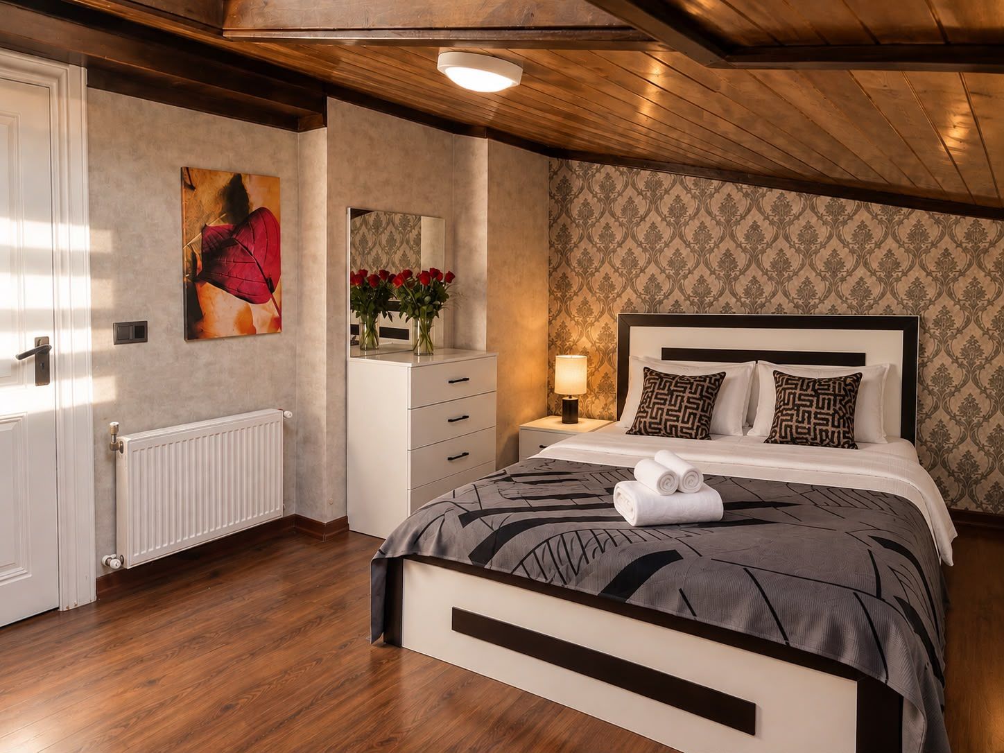 Un dormitorio con cama, mesita de noche, lámpara y cómoda con espejo y flores.