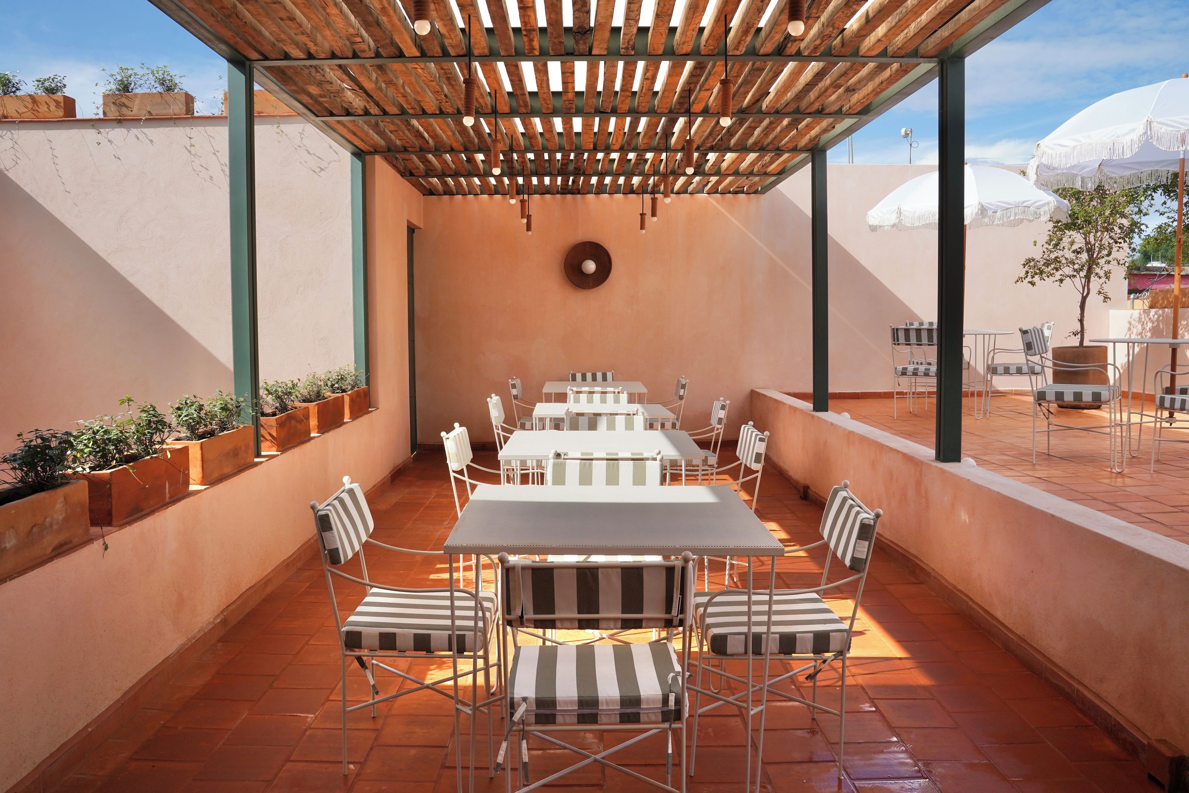 Une terrasse extérieure agrémentée d’une pergola en bois, de tables et de chaises en métal blanc, et de plantes en pot disposées le long des murs.