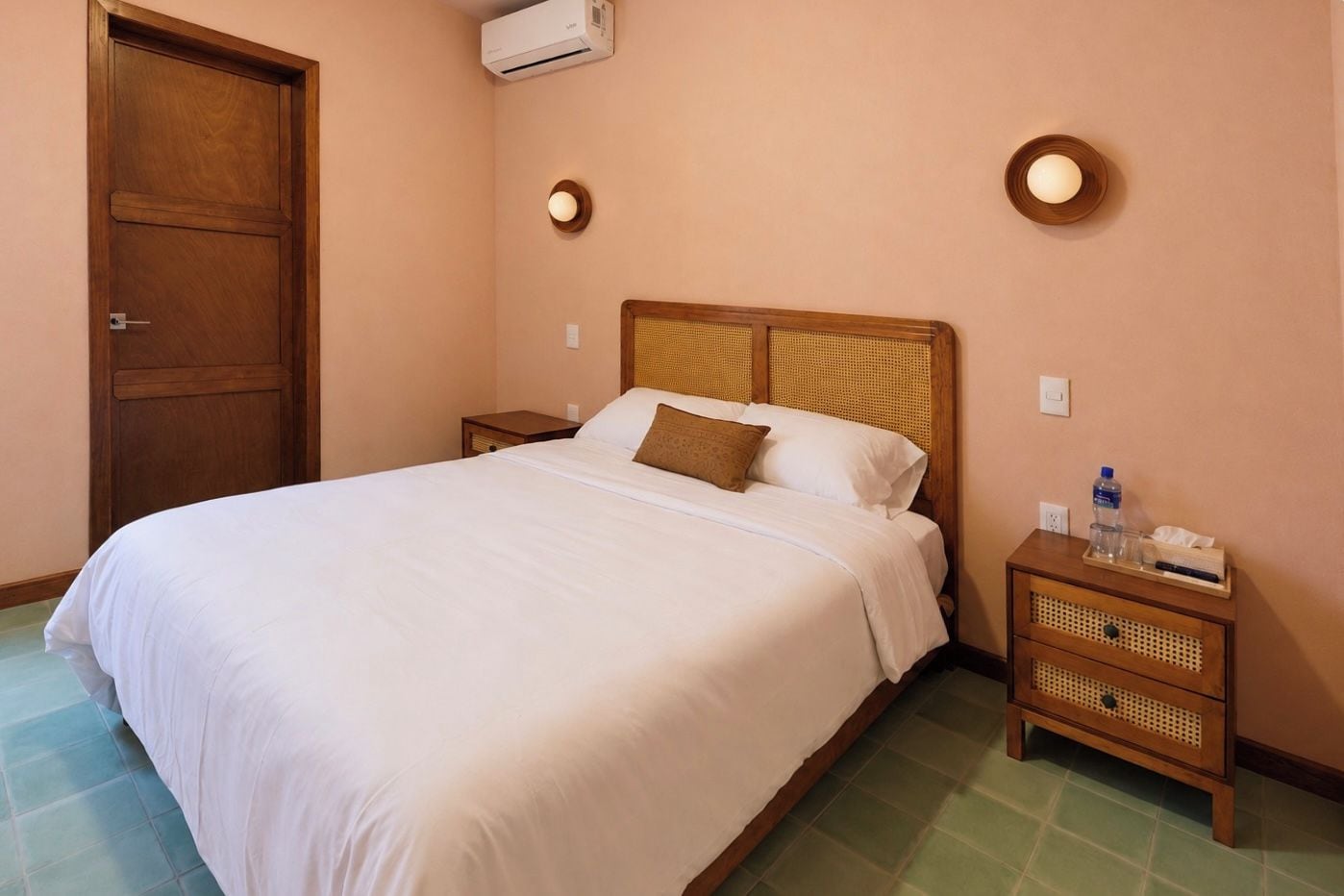 A hotel room with a bed, a nightstand, a wooden door, and an air conditioning unit.