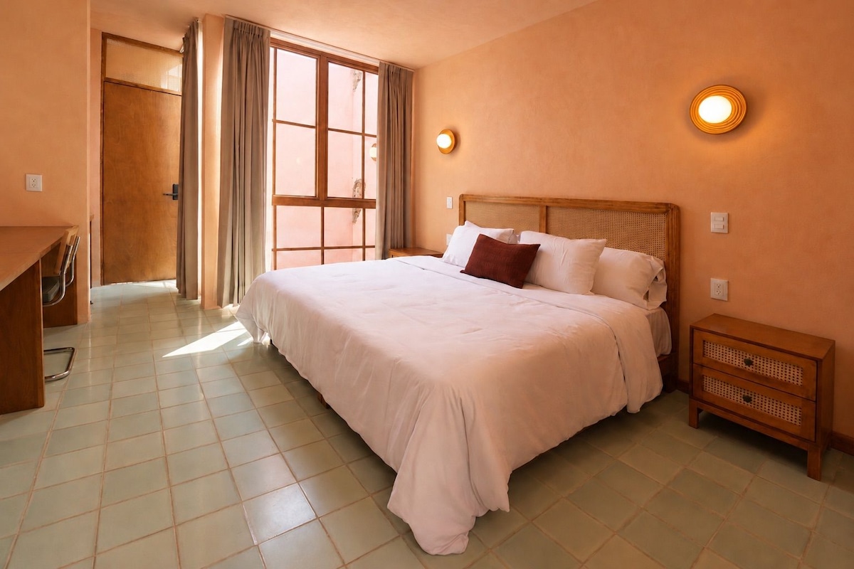 Une chambre à coucher avec un grand lit, une table de chevet, un bureau et une fenêtre avec des rideaux.