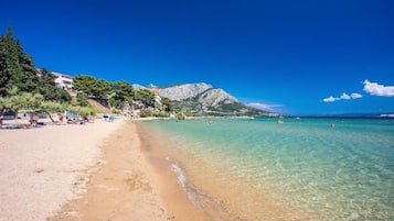 A beach with clear water, a mountain in the background, and buildings on the left.