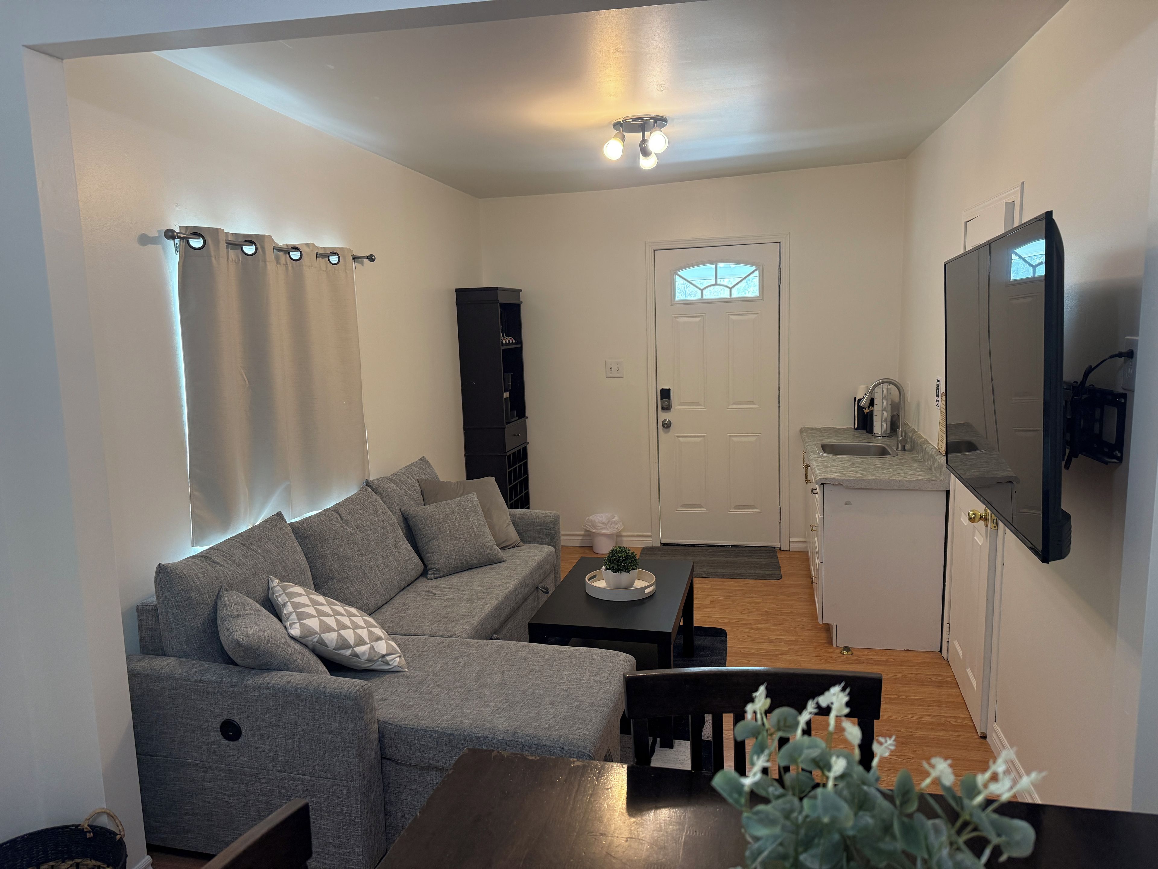 Uma sala de estar com um sofá cinzento, uma mesa de centro e uma mesa de jantar com cadeiras. Há uma área de cozinha com uma pia e um espelho.