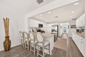 A modern kitchen with white cabinetry, a central island with bar stools, and a large window providing natural light. - Charming Fenced Eastport Cottage with Golf Cart (Wildwood)