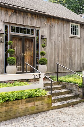 A rustic wooden building with a large wooden door, a set of stairs leading to a white porch with the word 'ONE' on it, and a small garden area with plants. - Stunning Post & Beam Cottage Located in the Heart of Essex Village (Essex)