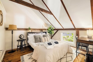 A bedroom with a sloped ceiling, a bed with pillows, a bedside table with a lamp, and a window with a view. - Stunning Post & Beam Cottage Located in the Heart of Essex Village (Essex)