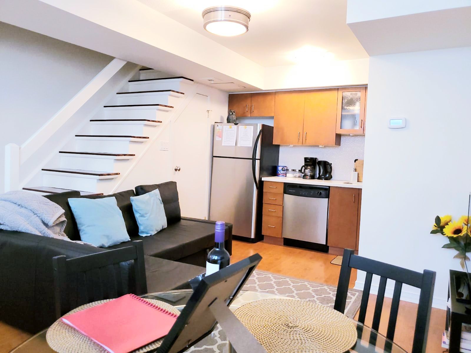 A compact living space with a staircase, a kitchenette, a dining area with a table and chairs, and a sofa with cushions.