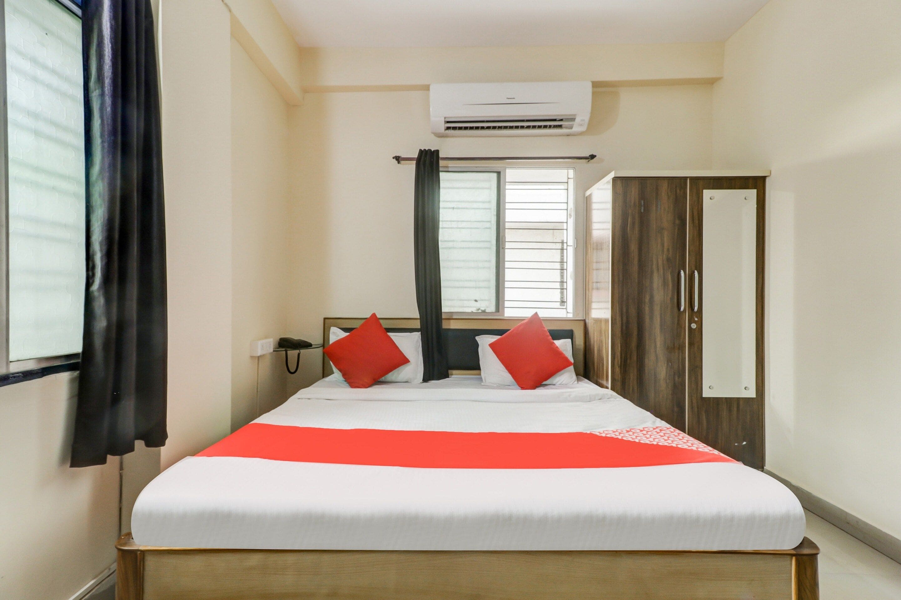 A hotel room with a bed, a wardrobe, an air conditioning unit, a window with curtains, and a bedside telephone.