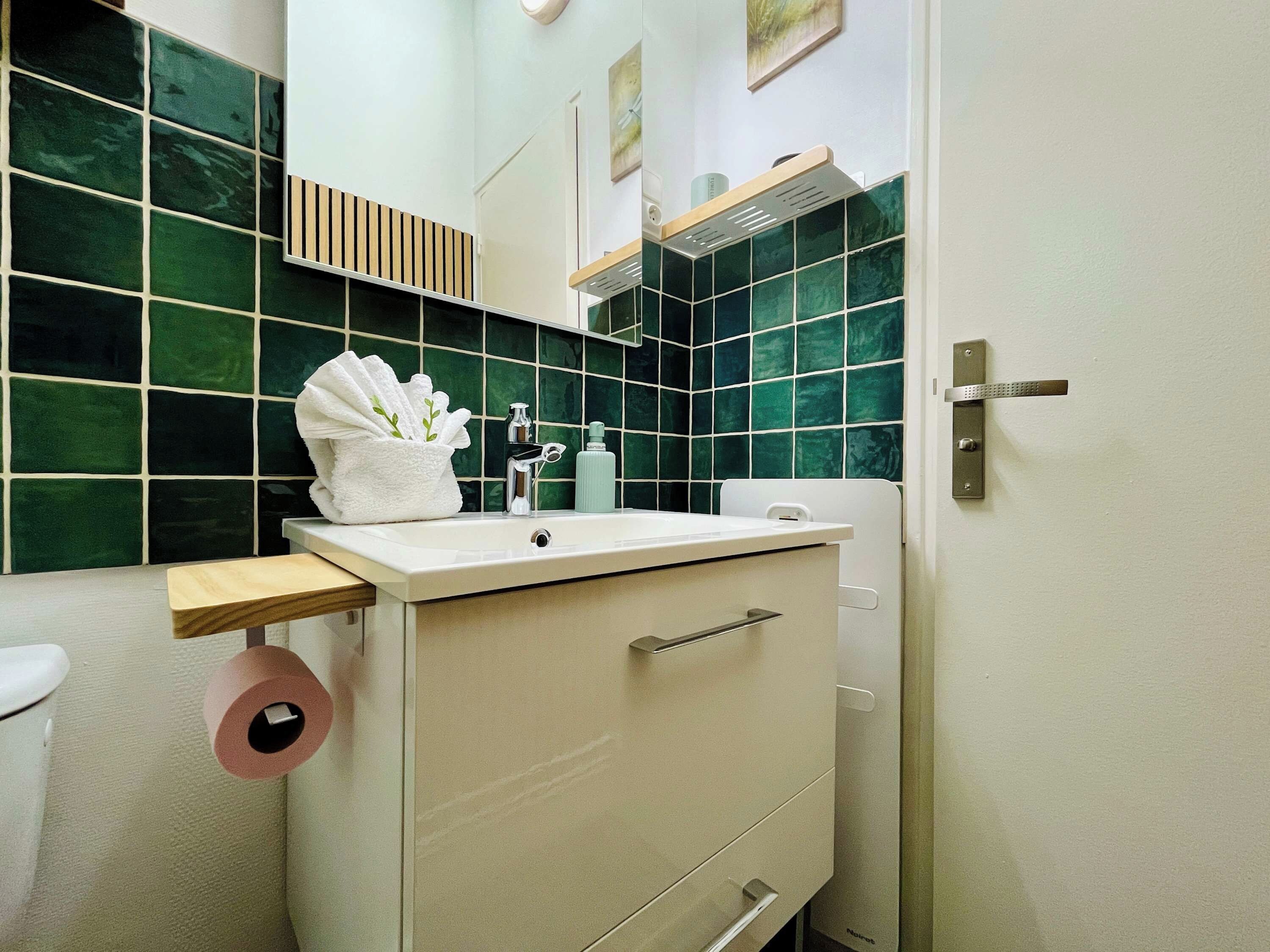 Kylpyhuoneessa on valkoinen pesuallas, valkoinen pyyhe pyyhetelineessä, wc-paperirulla ja vihreillä laatoilla päällystetty seinä.