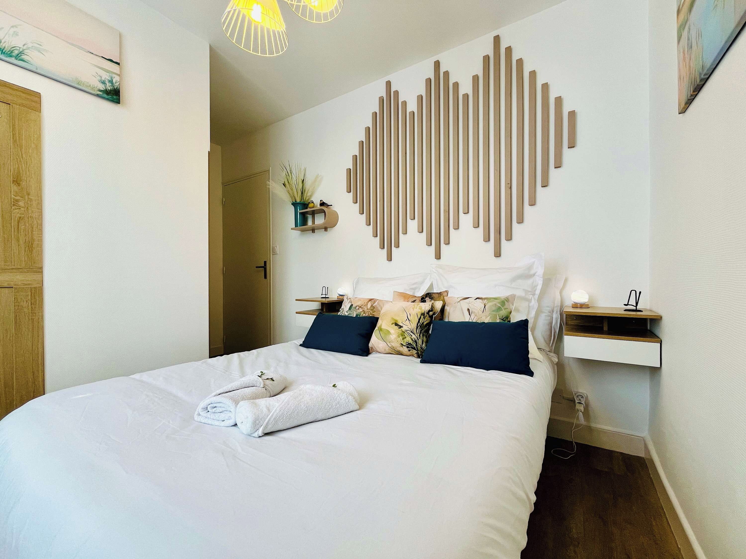 Una cama bien hecha con edredón blanco y toallas enrolladas, una mesita de noche con lámpara, y una lámpara de pared.