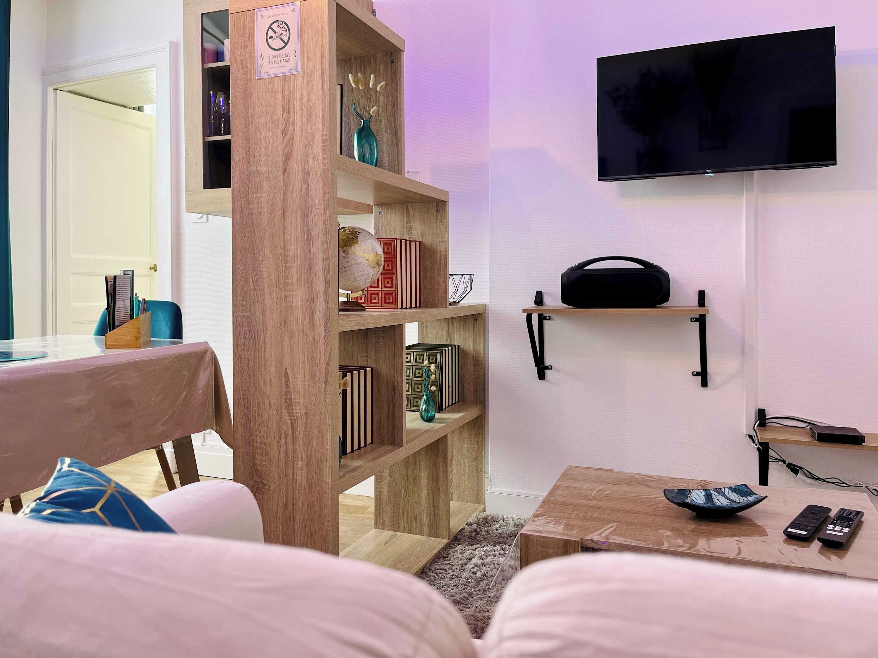 Ein Wohnzimmer mit einer Couch, einem Couchtisch, einem Bücherregal, einem an der Wand befestigten Fernseher und einer kleinen Ablage mit einem Lautsprecher.