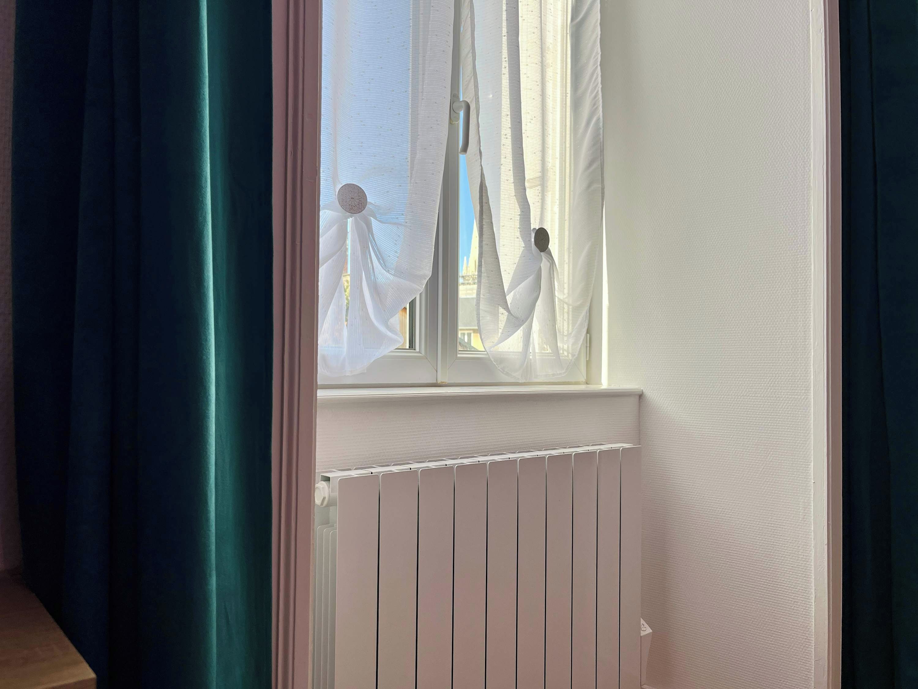 Une fenêtre avec des rideaux légers, un radiateur blanc en dessous, et une vue sur l’extérieur.