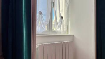 Une fenêtre avec des rideaux légers, un radiateur blanc en dessous, et une vue sur l’extérieur.