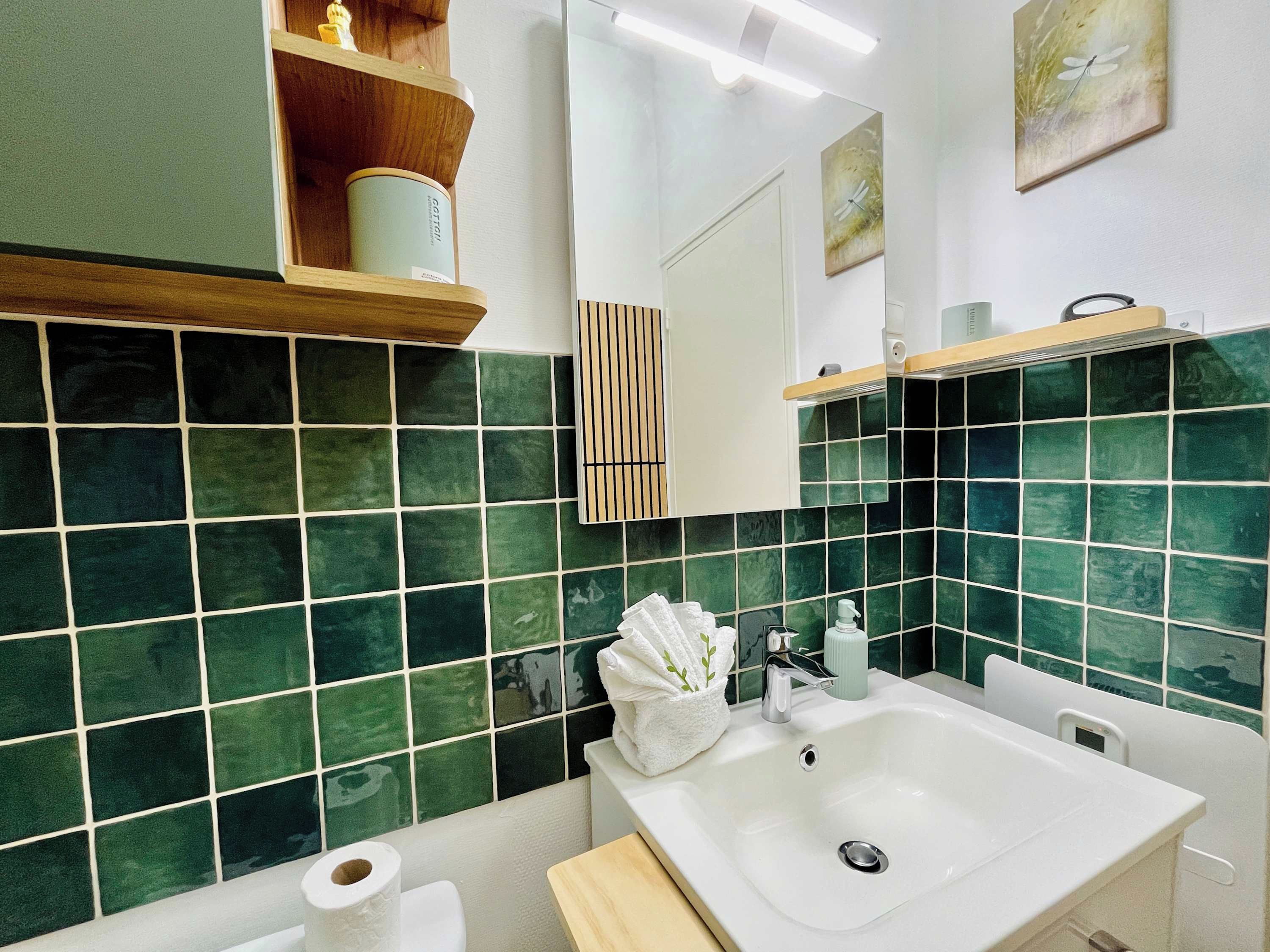 Une salle de bain avec des carreaux verts, un lavabo blanc, un miroir et une étagère contenant des articles de toilette.