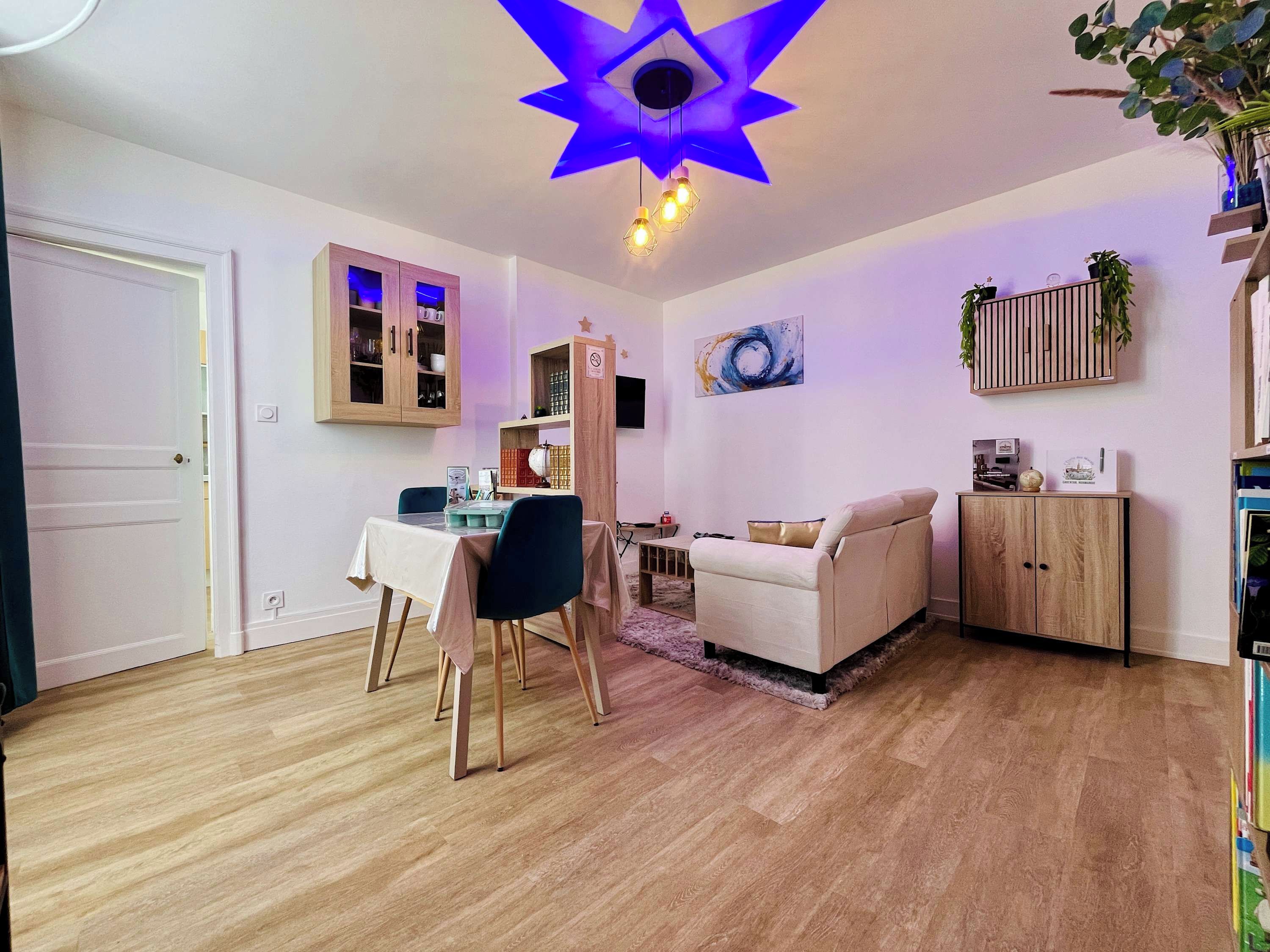 Ein Wohnzimmer mit Essbereich, Holzboden, einer Deckenleuchte und einem an der Wand befestigten Regal mit Pflanzen.