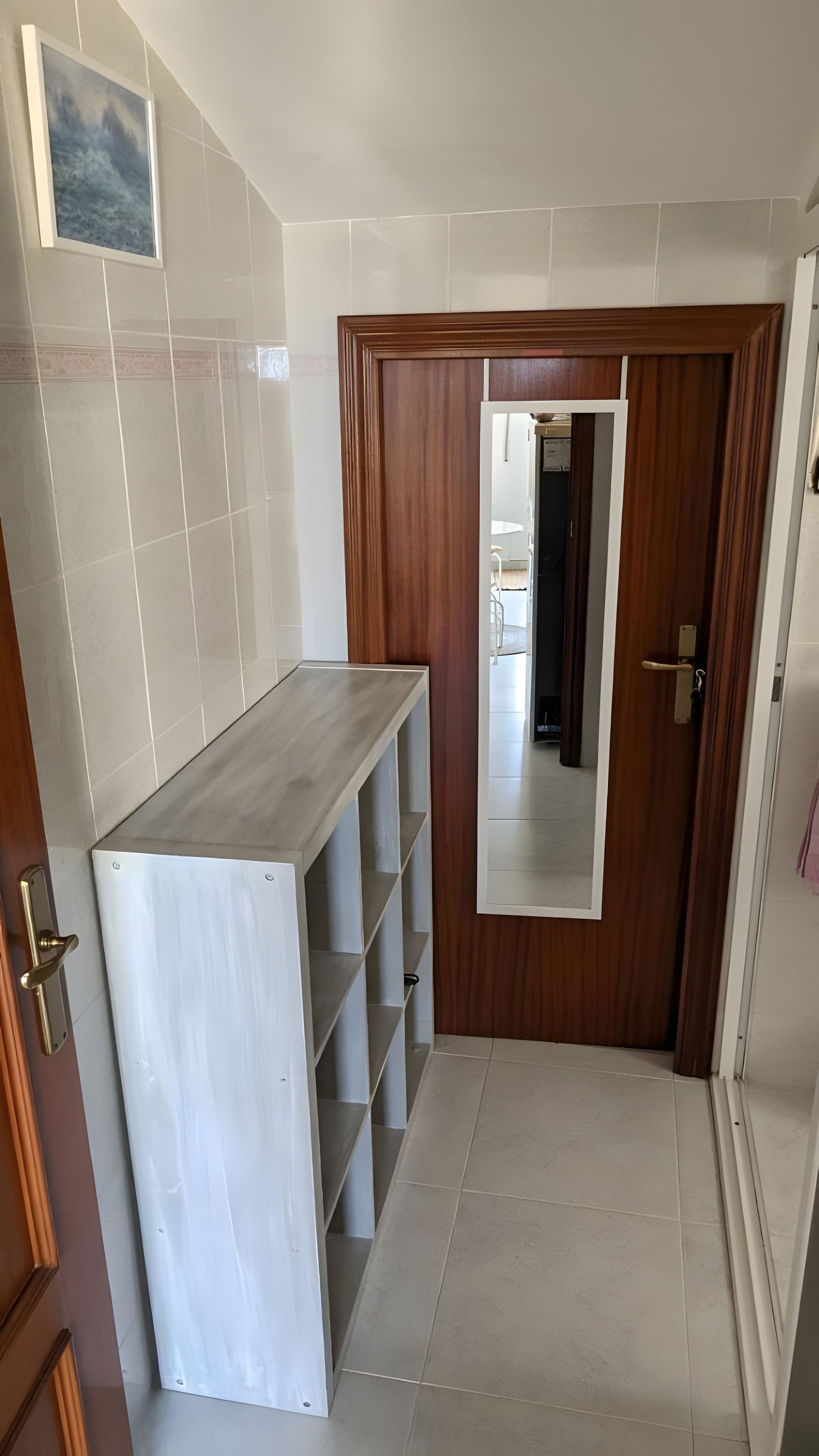 Un couloir avec un sol carrelé de carreaux blancs, une porte en bois et un meuble blanc avec des étagères.