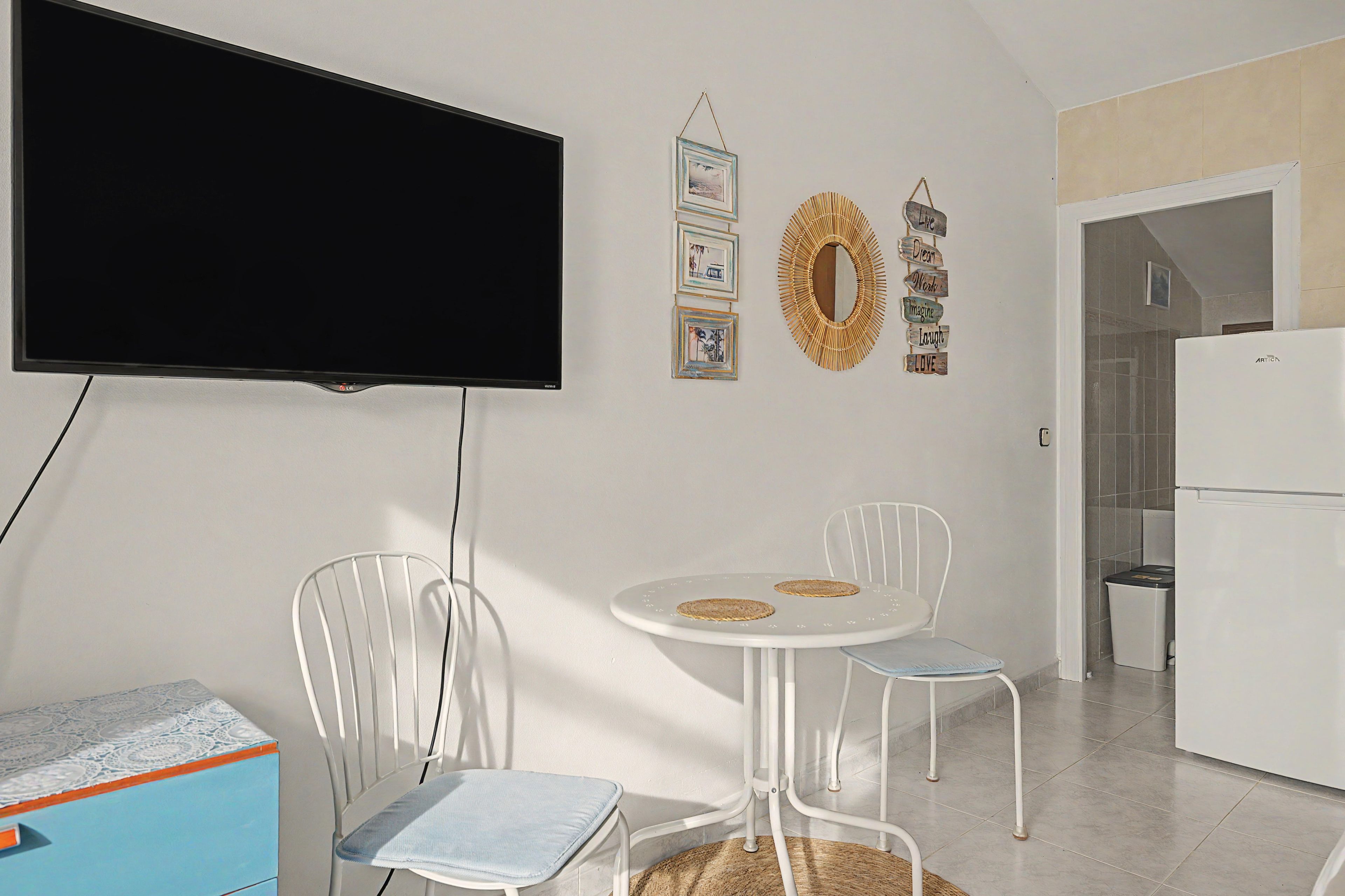 A modern kitchen with a flat-screen TV, a dining table with chairs, and a refrigerator.
