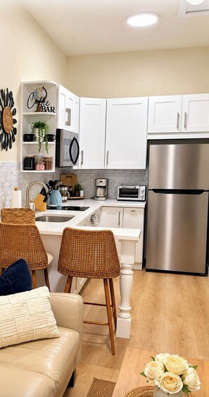 Een moderne keuken met witte kasten, een roestvrijstalen koelkast, een koffiehoek en een eethoek met rieten stoelen en een bank.