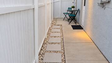 A pathway leading to a patio area with a white picket fence, outdoor seating, and a building with a light fixture.