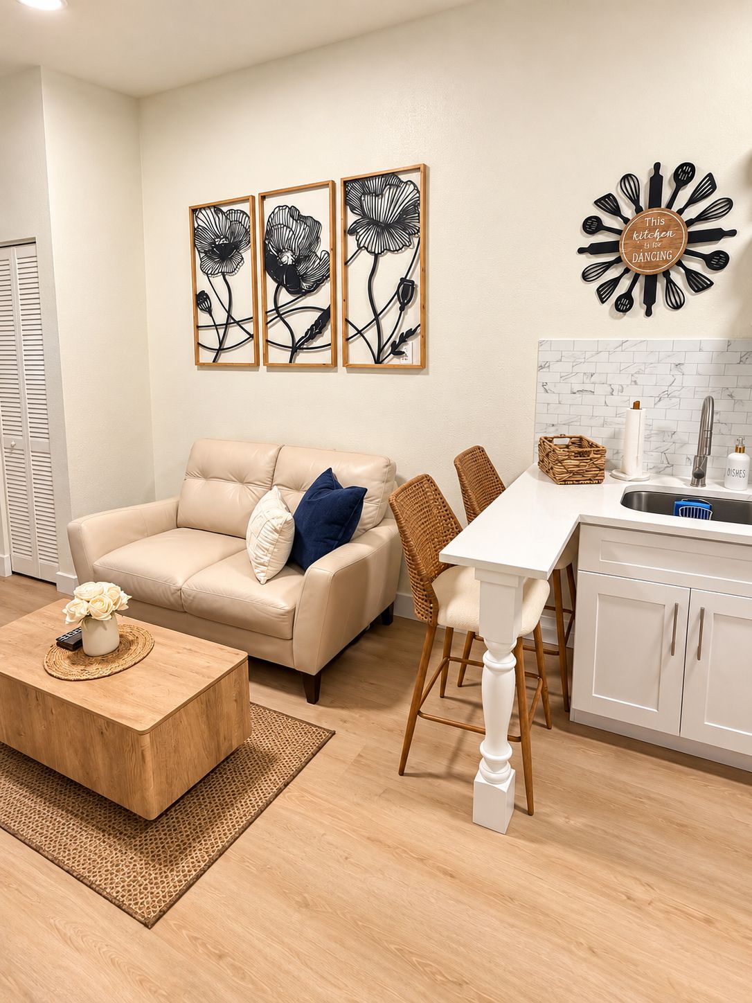 A modern living room with a beige sofa, a coffee table with a vase, a dining area with a white table and wicker chairs, and a kitchen with a white countertop and a backsplash.