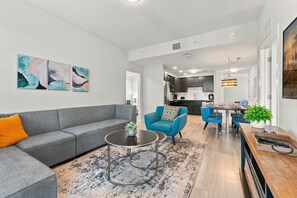 A modern living room with a grey sofa, blue armchairs, and a coffee table. There is a kitchen in the background with dark cabinets and a dining area with a wooden table and chairs. - Stay Central, Explore More, Orlando 3BR Condo (Orlando)