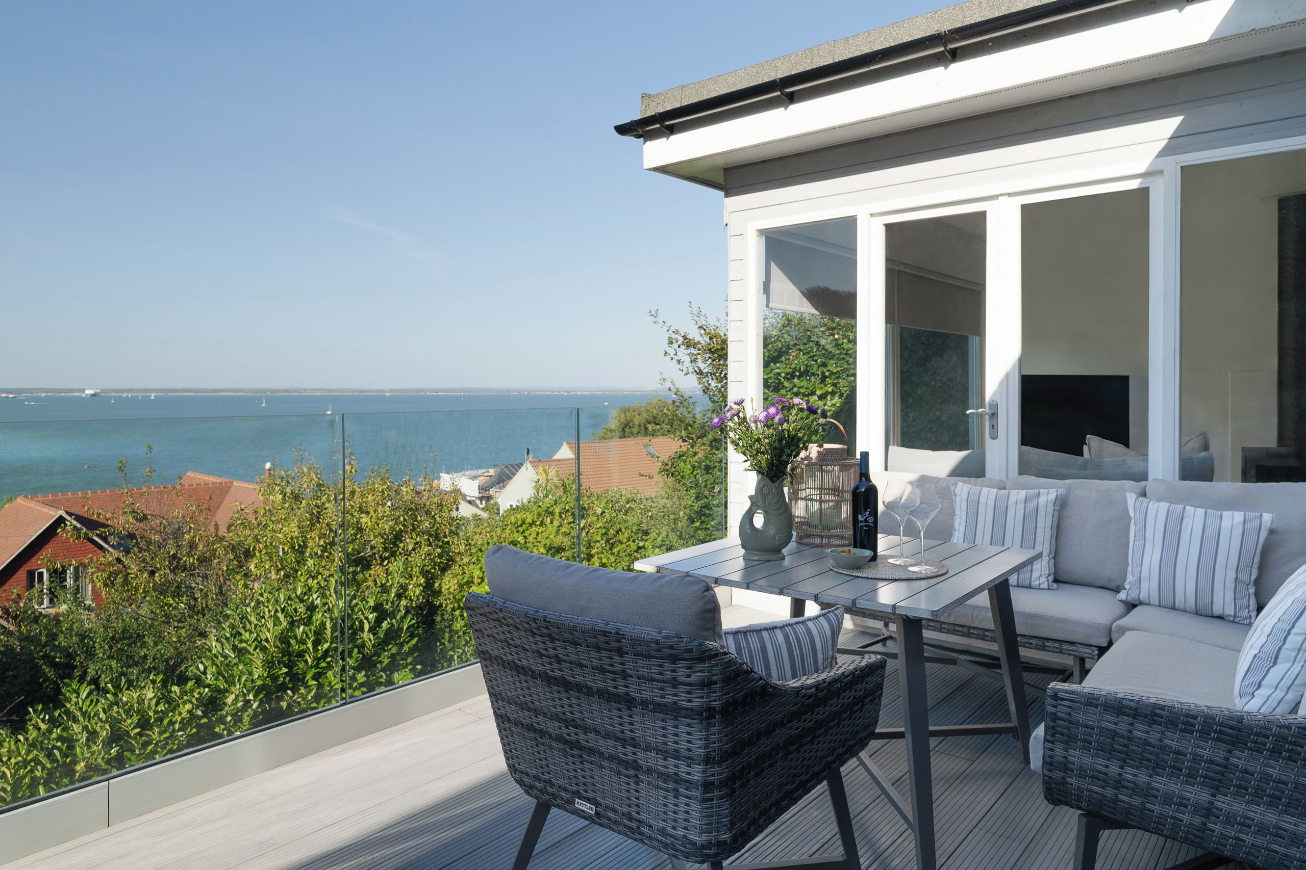 A balcony with a view of the sea, wicker furniture, and a table with a vase and wine glasses.