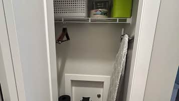 A narrow hallway with a door leading to a closet containing shelves with storage baskets, a green container, and a roll of toilet paper.