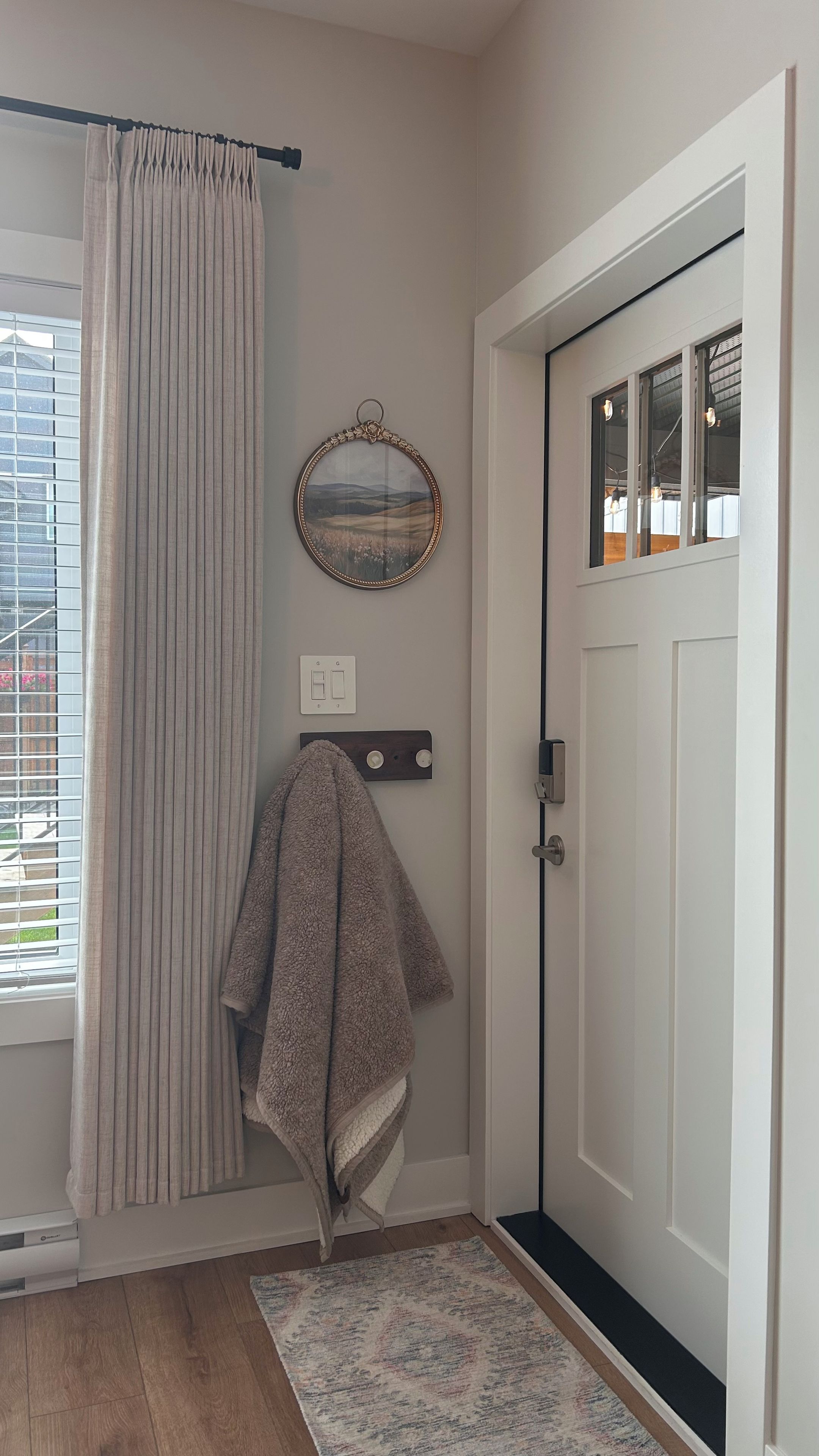 Ein Zimmer mit einer weißen Tür, einem Fenster mit Jalousien, einem an der Wand hängenden Bilderrahmen, einem Handtuch an einem Haken und einem gemusterten Teppich auf dem Boden.