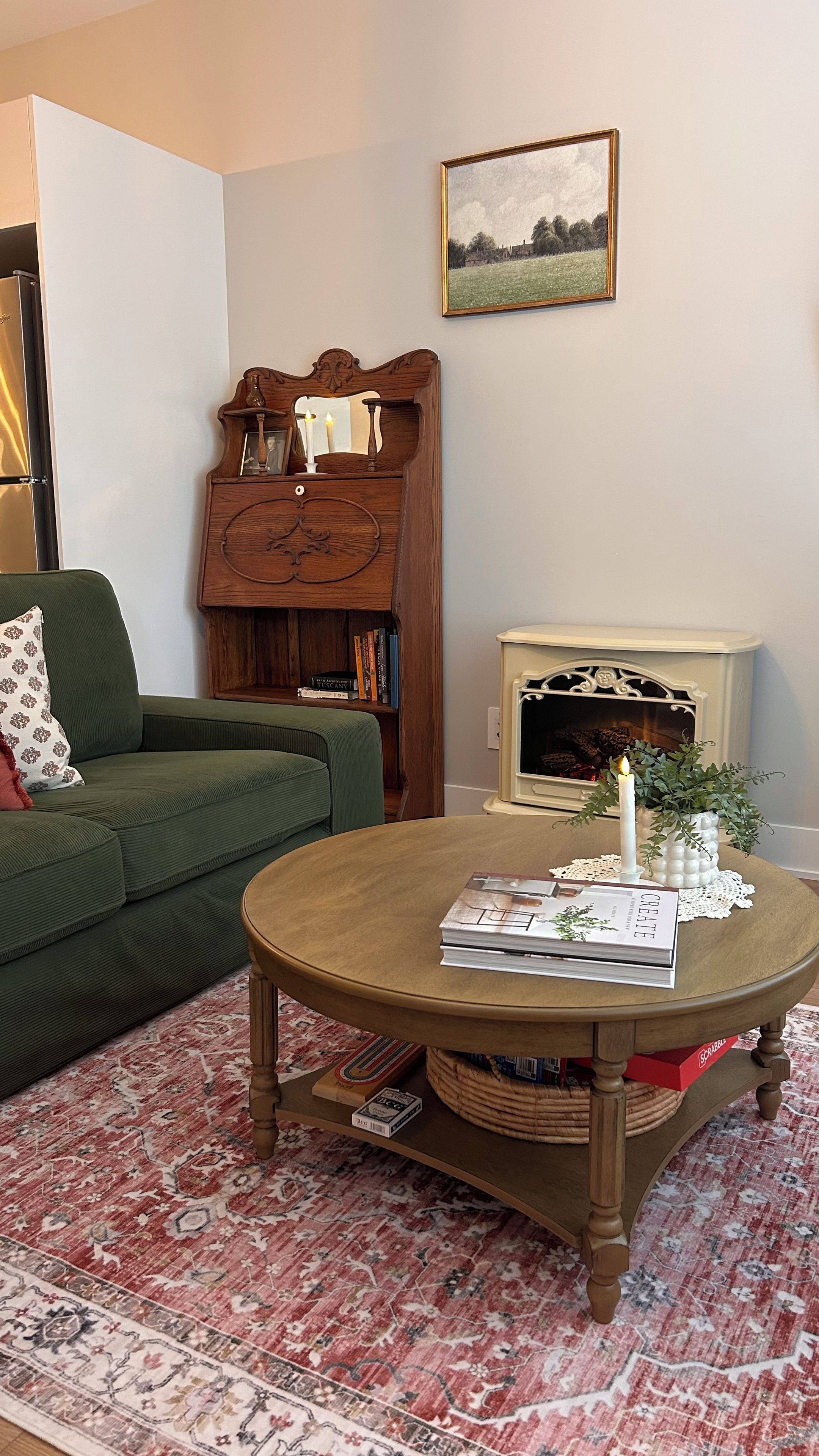 Una sala de estar con un sofá verde, una mesa de centro de madera con libros y una vela, una chimenea y un cuadro de paisaje en la pared.