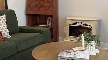Una sala de estar con un sofá verde, una mesa de centro de madera con libros y una vela, una chimenea y un cuadro de paisaje en la pared.