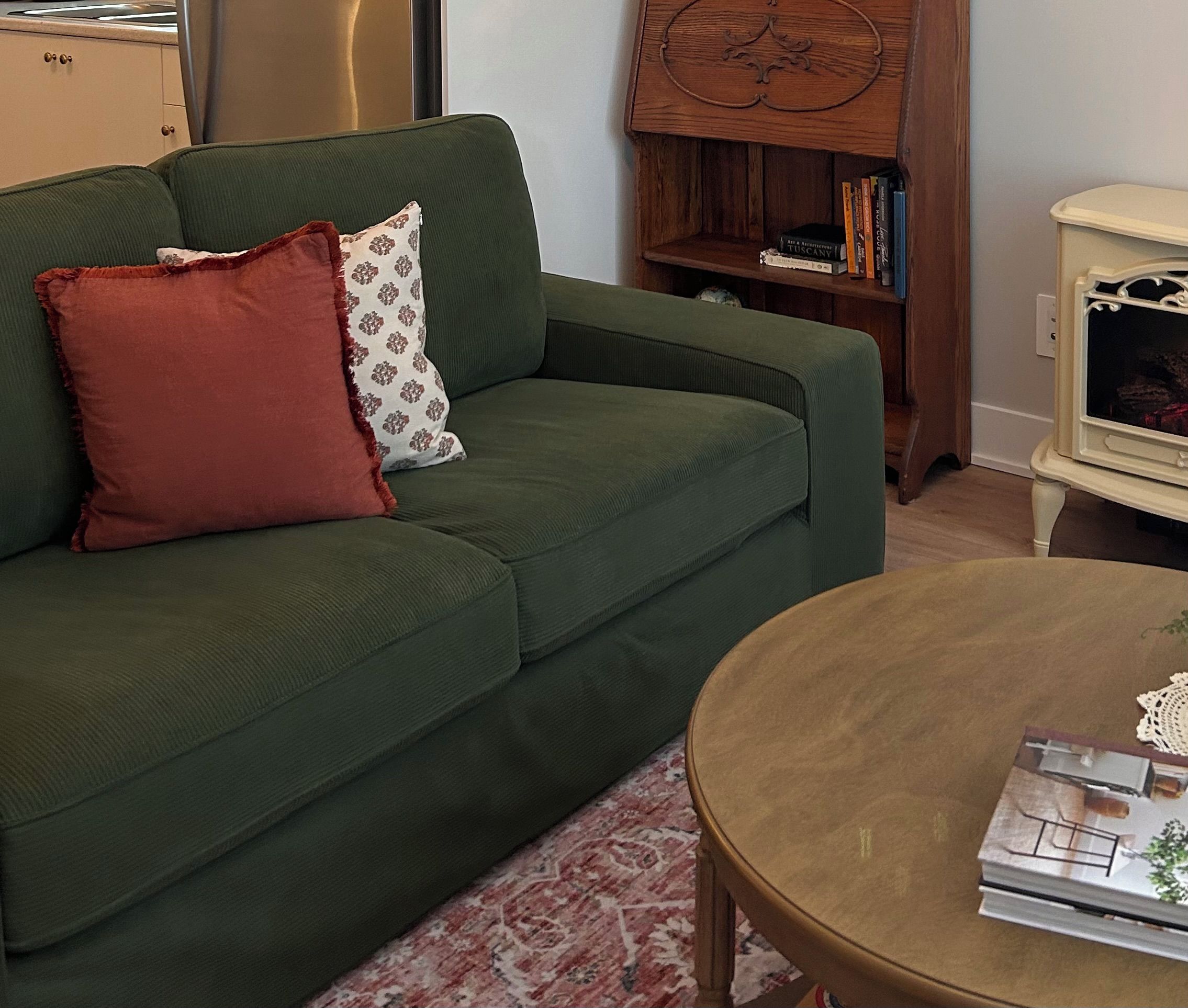 Un canapé vert avec deux coussins, une bibliothèque en bois remplie de livres, une table ronde avec un magazine, et une petite armoire blanche avec un miroir.